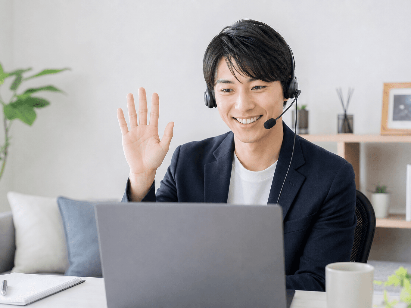 ヘッドセットを装着して画面越しの相手へ笑顔で手を振る在宅ワーク中の男性会社員