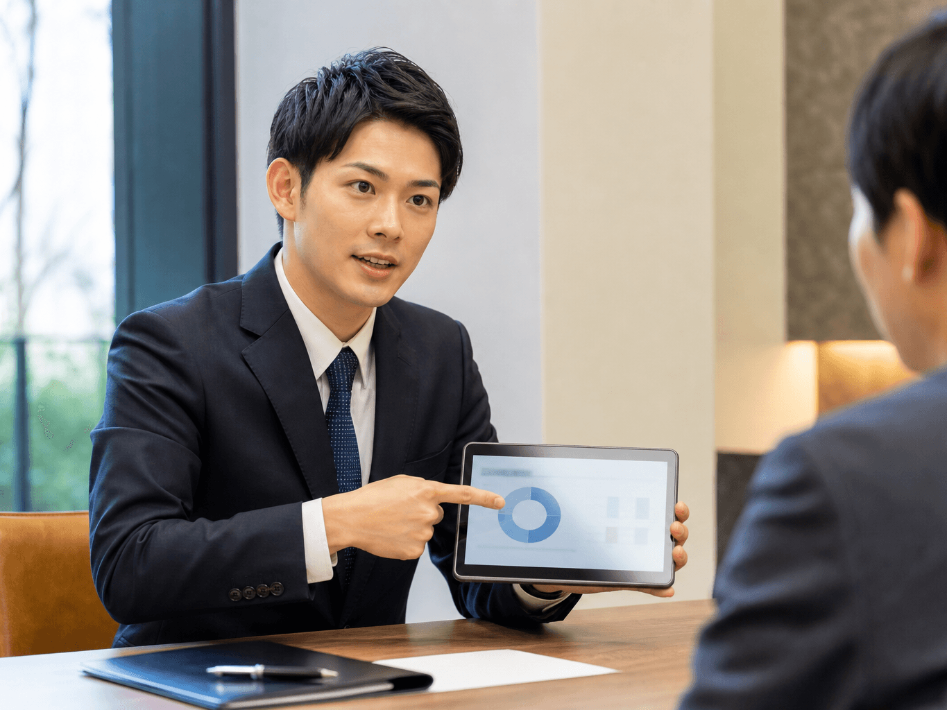 商談席でタブレットの資料を指差しながら顧客へ熱心に説明する男性営業担当者の様子